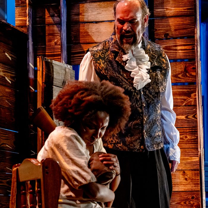 A man in a brocade waistcoat standing over a young woman seated in the cabin